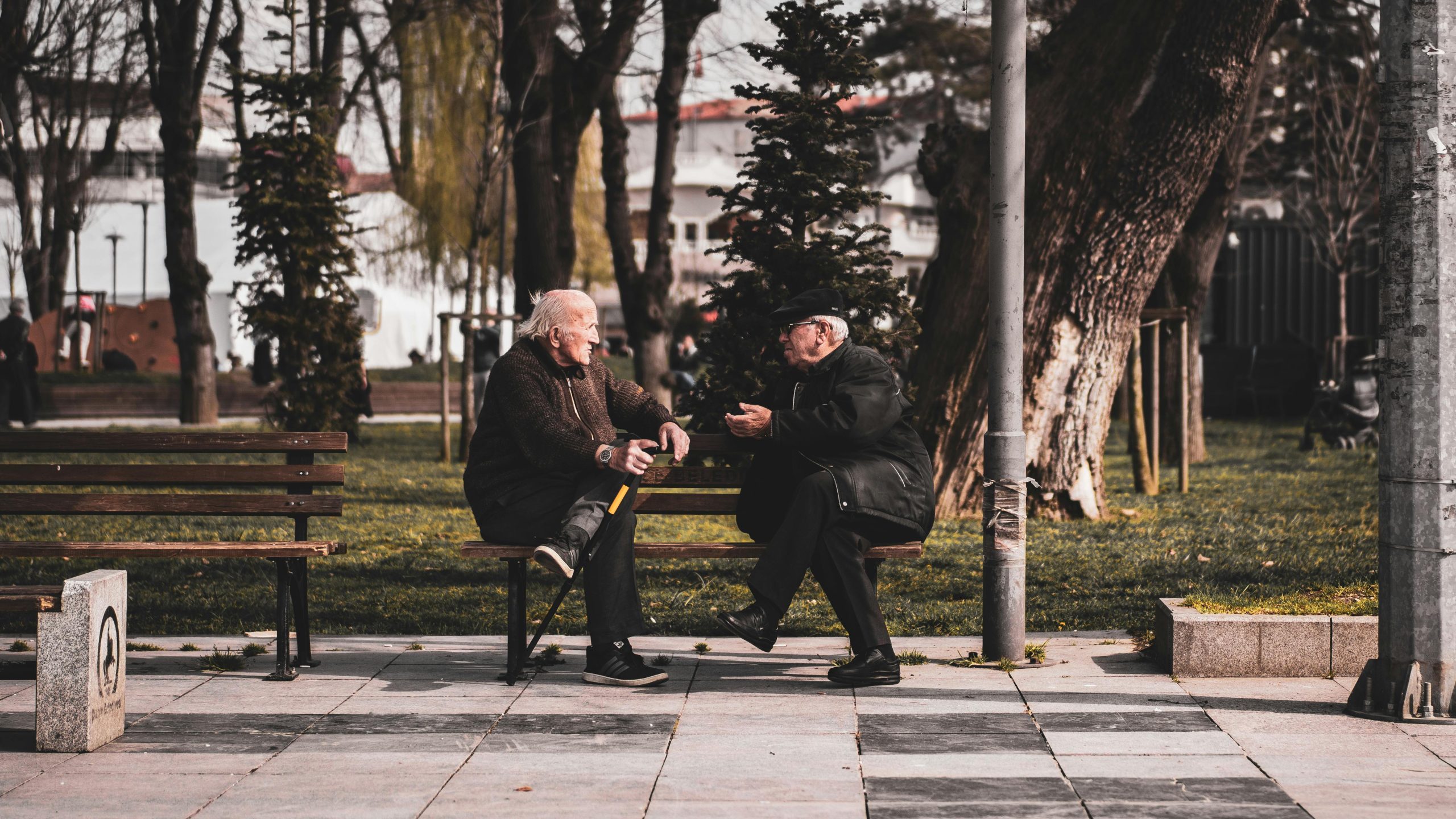 two elderly men wearing coats and hats, sitting on a park bench, talking to each other